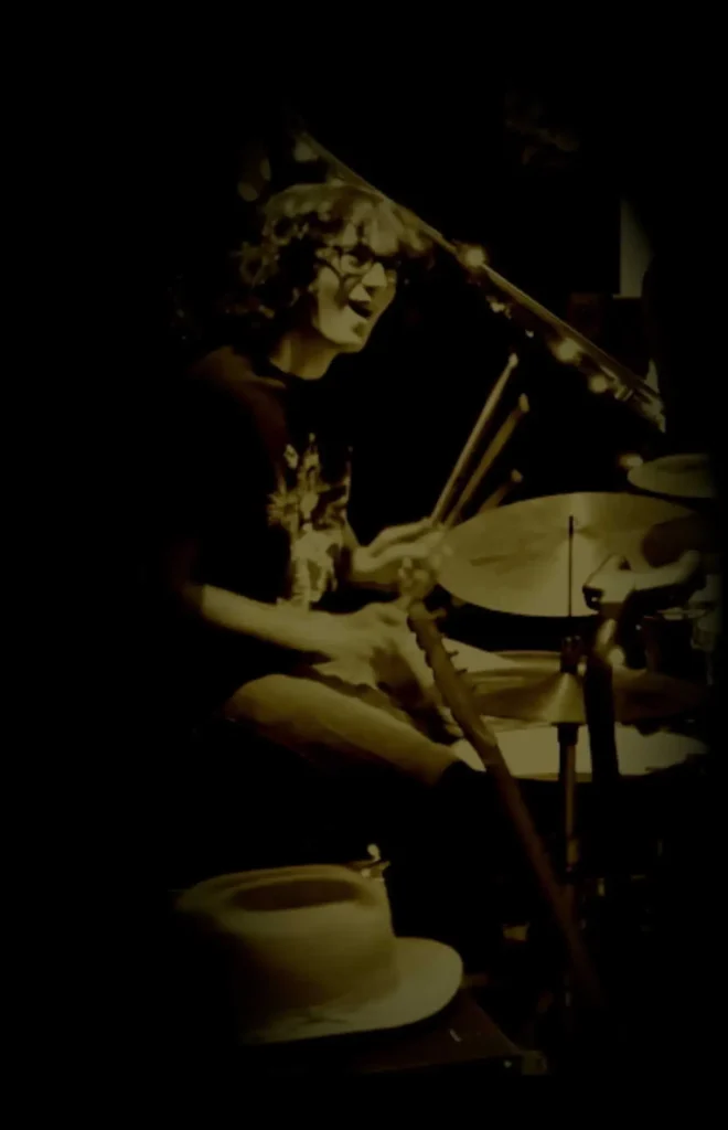 - Dante Kierstead Drummer playing a drum set on a dimly lit stage, with sticks in mid-motion in a sepia-toned scene.
